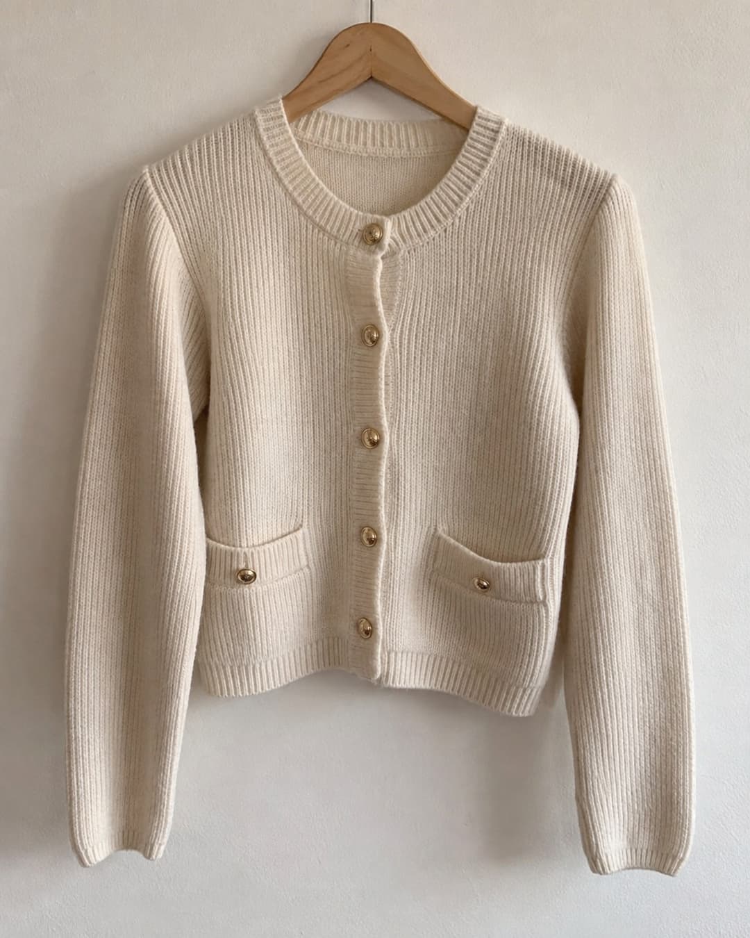 A cream knit cardigan with gold buttons on a wooden hanger against a plain wall
